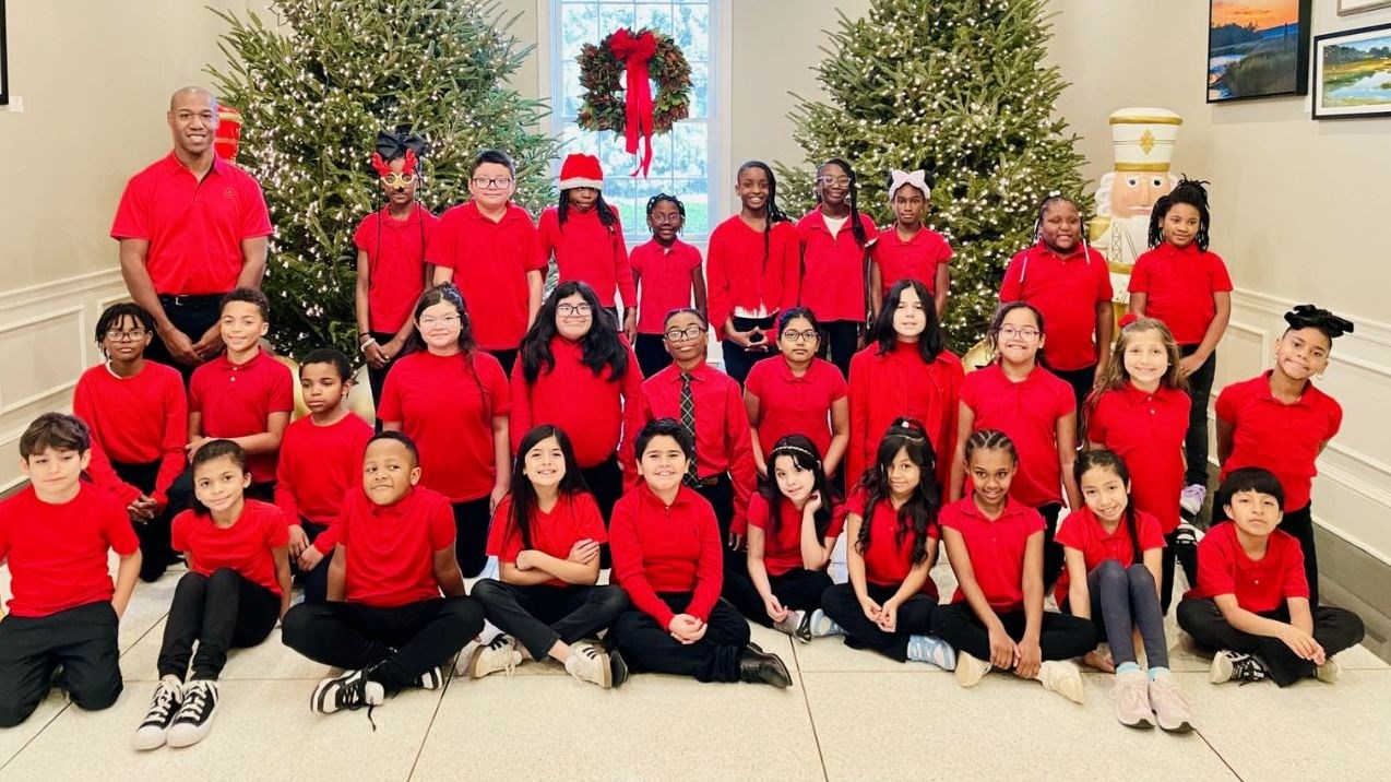 Choral director and choral students posing for picture in front of two Christmas trees. Each person is wearing a red shirt.
